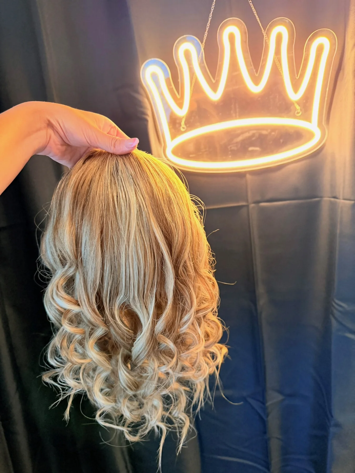 Honey-blonde curled ponytail hairpiece held beneath a glowing crown sign