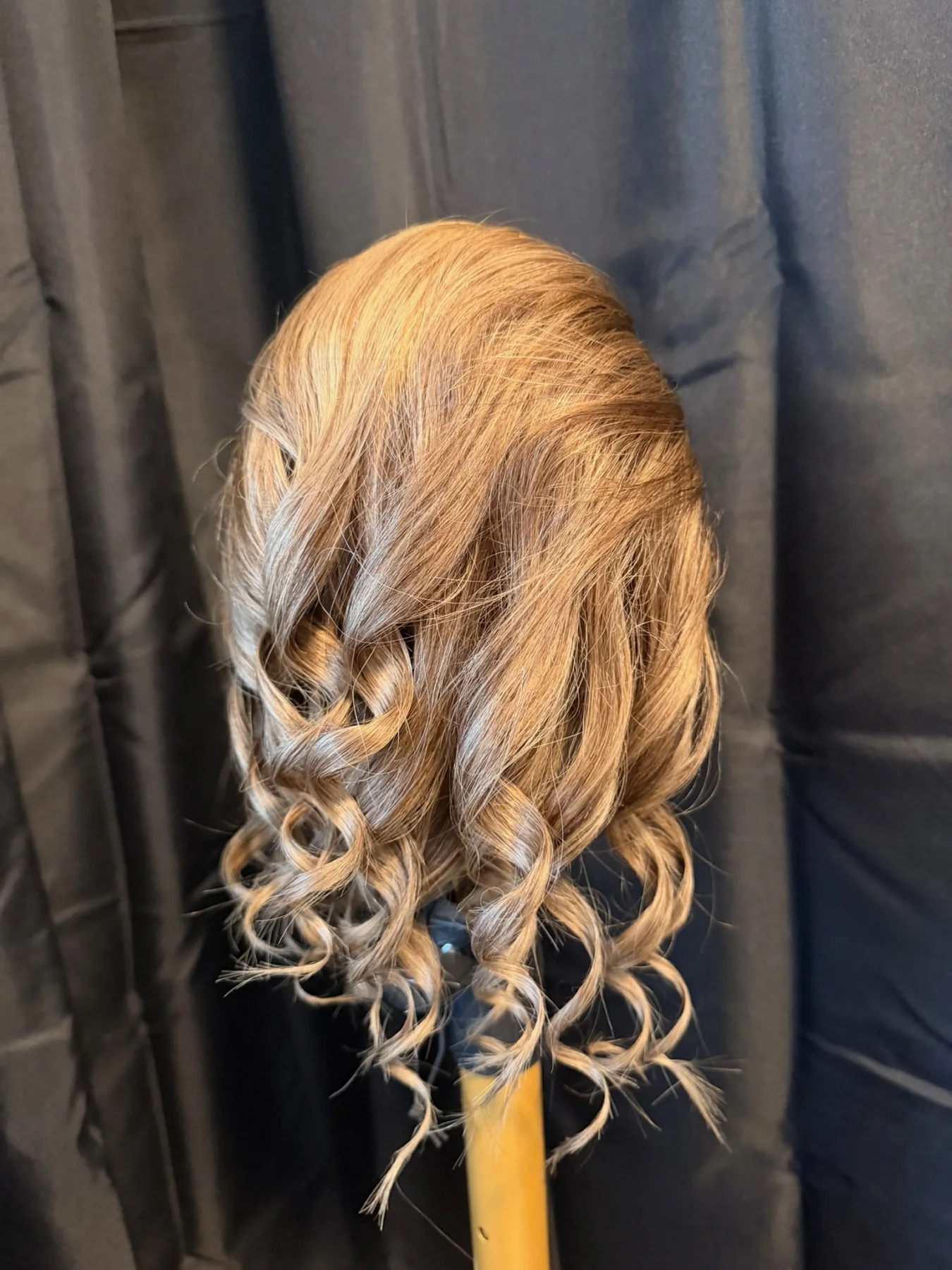 Light ash-brown ponytail hairpiece with curled ends displayed on a stand against black