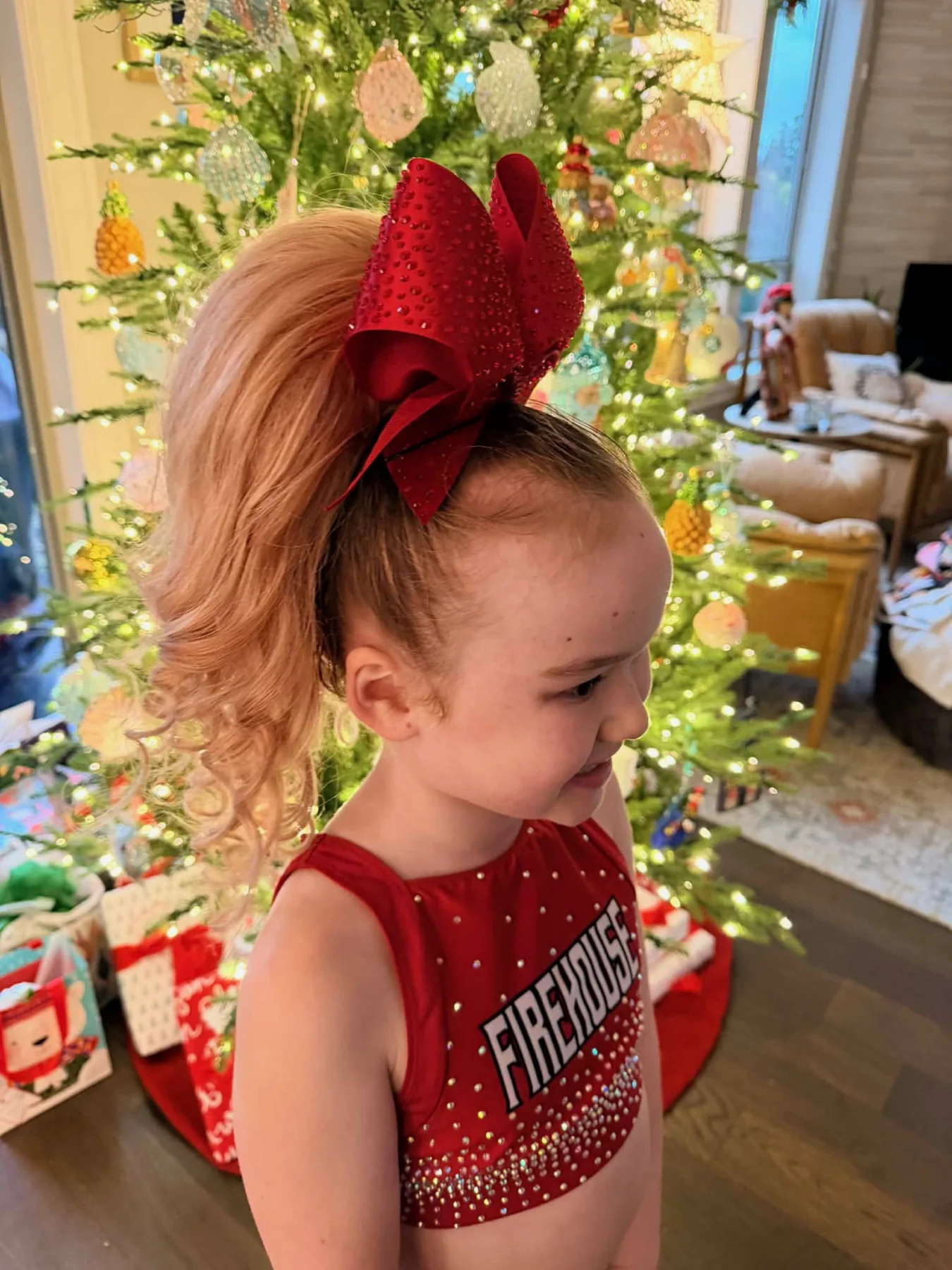 Young cheerleader wearing a curled strawberry-blonde ponytail and red rhinestone bow beside a Christmas tree