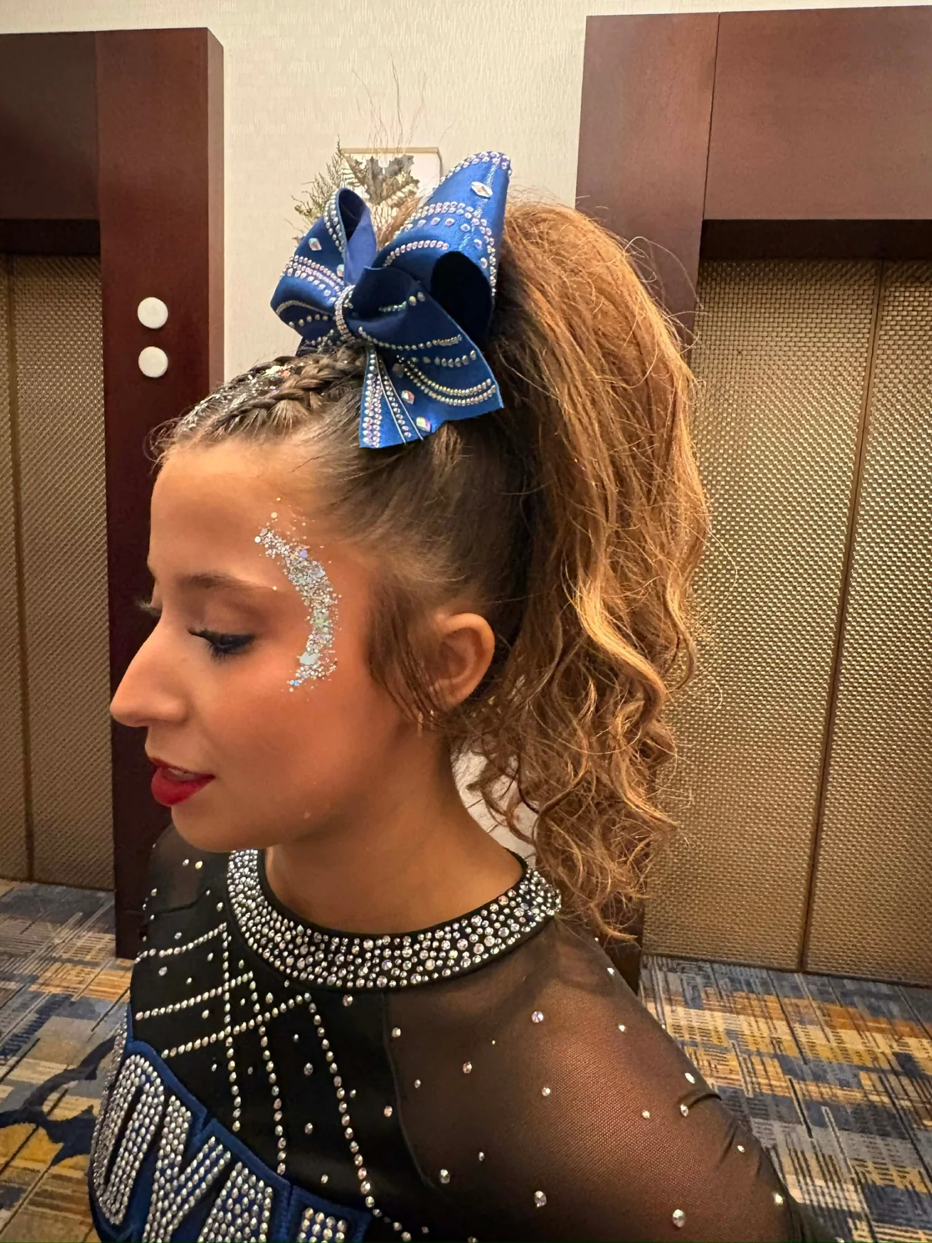 Cheerleader in profile wearing a long curled brown ponytail and royal-blue rhinestone bow
