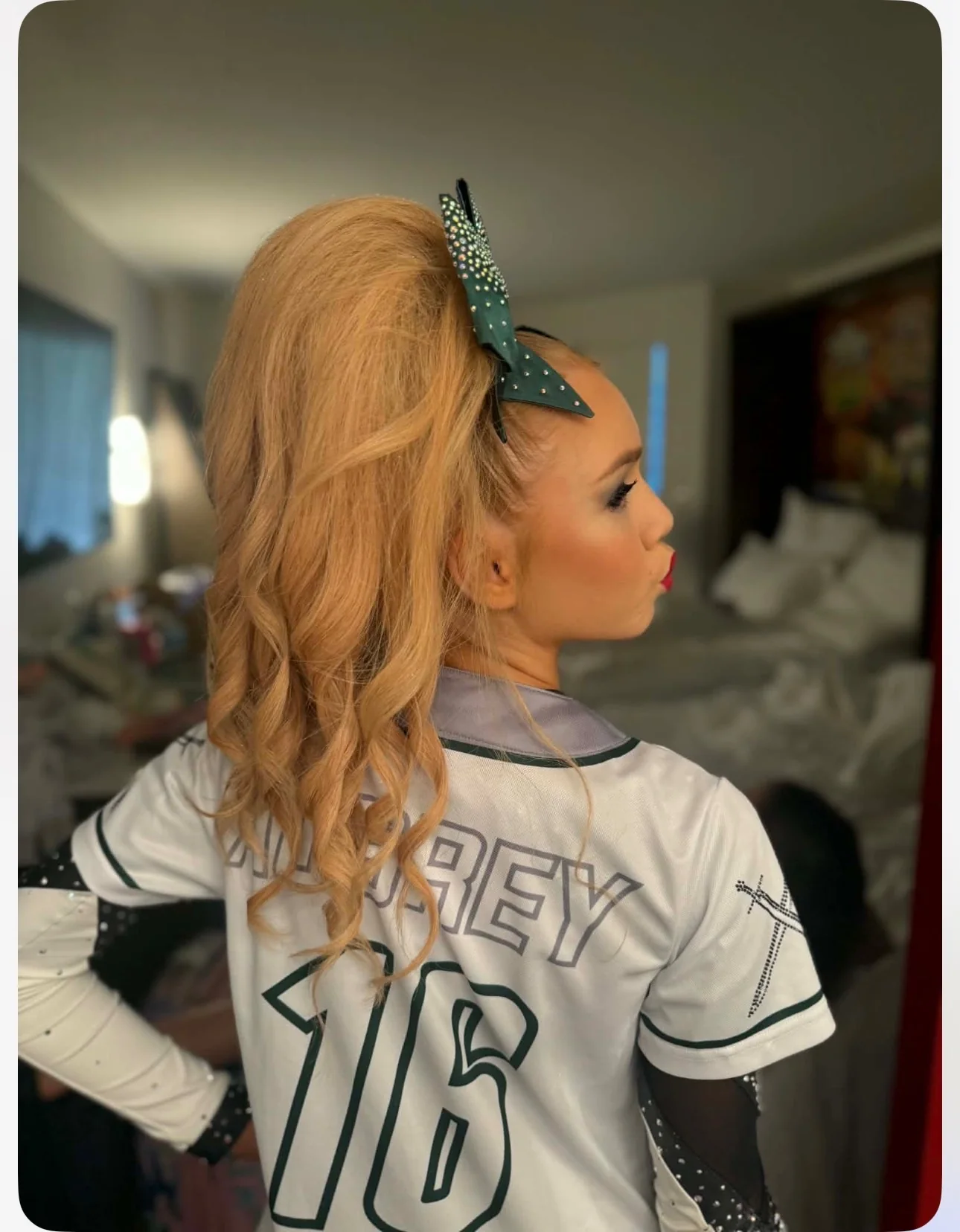 Side view of a cheerleader wearing a long blonde wavy ponytail and dark-green rhinestone bow