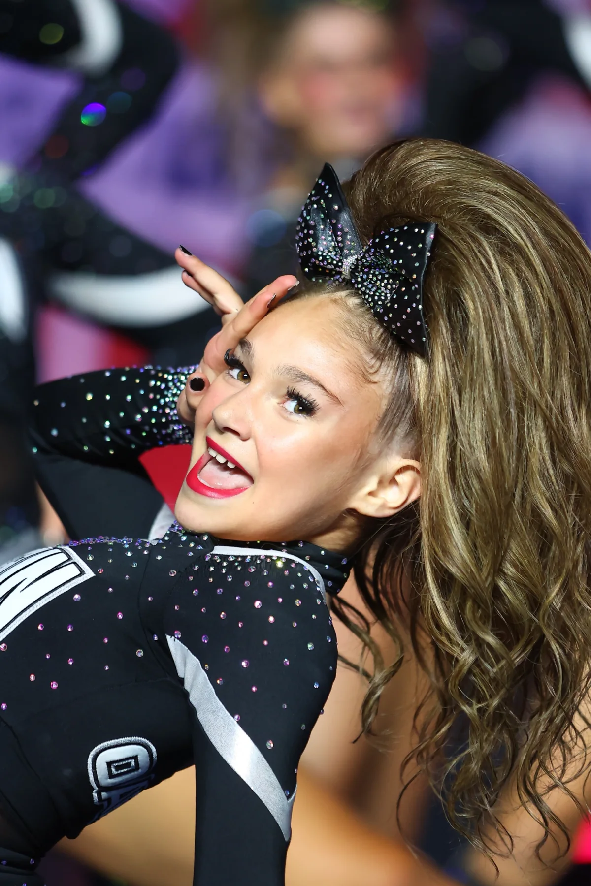Cheerleader smiling during a routine with a highlighted brown high ponytail and black rhinestone bow