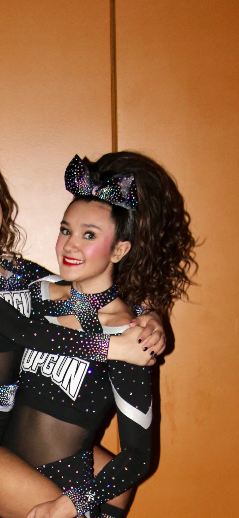 Top Gun cheerleader posing in front of an orange wall with a dark curly ponytail and black rhinestone bow