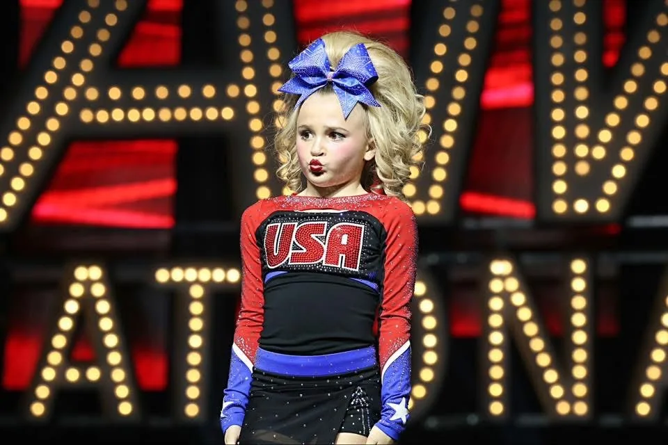 Young USA cheerleader on stage wearing a platinum-blonde curly ponytail and blue rhinestone bow