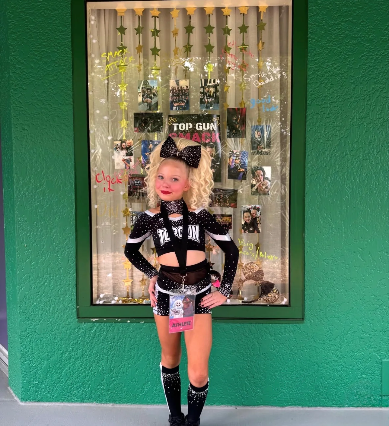 Young Top Gun cheerleader posing by a team display with a blonde curly ponytail and black rhinestone bow