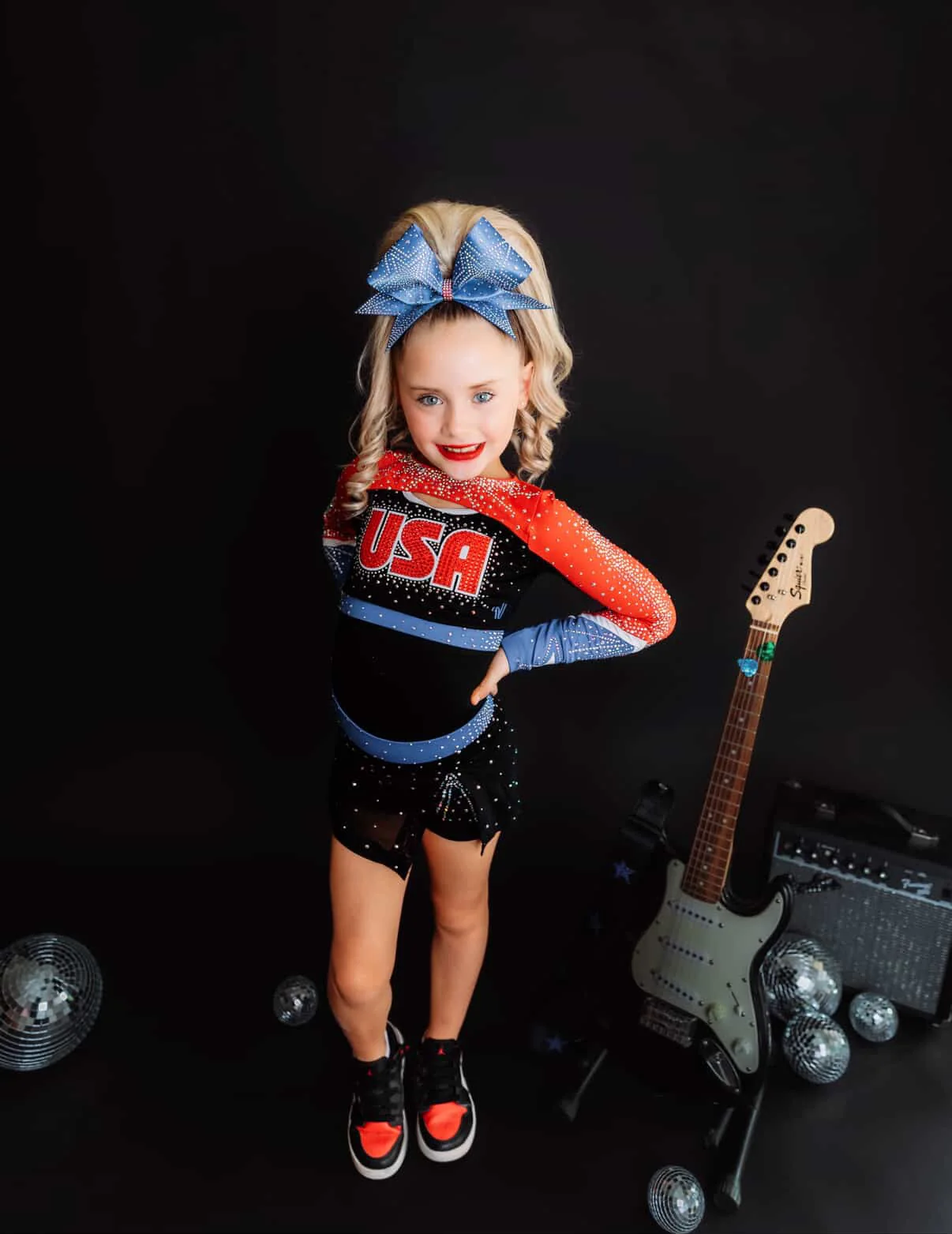 Young USA cheerleader posing in a studio with a blonde curly ponytail and blue rhinestone bow